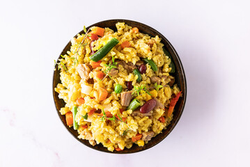 Stewed rice with meat and assortment on vegetables and spices in dark ceramic bowl on white marble table top view. Tasty homemade food concept.