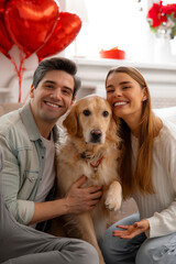 Smiling young couple posing with their dog at home, celebrating Valentine’s Day together. Cozy...