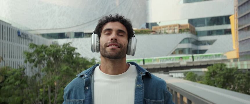 Portrait of curly haired Hispanic man puts on headphones and listens to music on street anamorphic lens. Latin guy in denim jacket feels happy as hears song in financial district