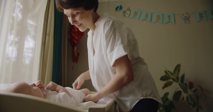 Mother changing diaper for her baby on bed. Happy infant comfortable when cleaning. Mother changing her baby's diaper playing with child. Mother changes baby white clean diaper at home on the bed.