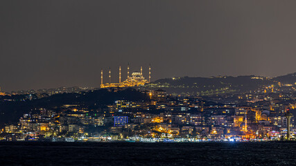 Panoramic view of Istanbul skyline with the camlica mosque rising above the city, ferries crossing the bosphorus strait, and dense urban architecture under dramatic clouds.