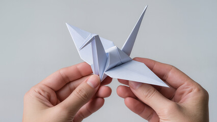 A close-up of female hands carefully holding a neatly folded white origami crane. The neutral light background emphasizes the clean lines and minimalism of the composition. The image symbolizes hope, 