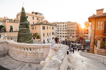 Woman with her dog enjoying a festive sunset moment overlooking Rome near the Spanish Steps with a decorated Christmas tree and lively square below. Holiday atmosphere, winter tourism and European