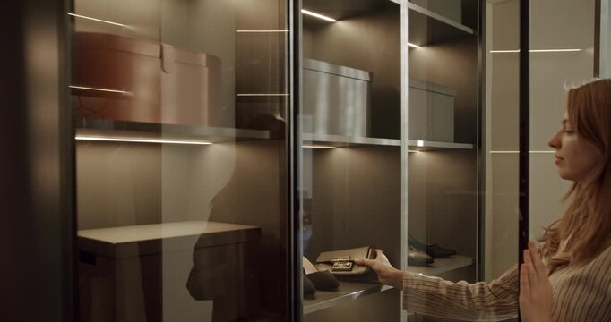 A young woman opens a modern wardrobe with glass doors, revealing organized shelves with shoes, bags, and storage boxes. The wardrobe is illuminated with built-in lighting,