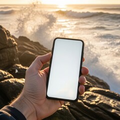 Hand holding a smartphone with a blank display against a beautiful coastal sunset with crashing waves