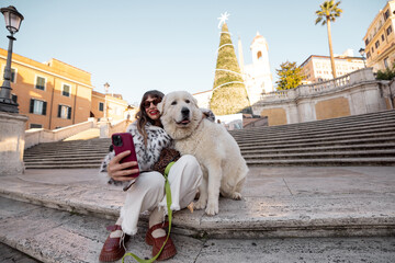 Woman taking a festive selfie with her dog near the Spanish Steps in Rome with a decorated Christmas tree in the background. Holiday atmosphere, winter tourism, joyful city break travel and European