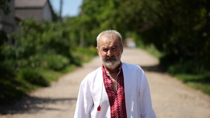 Elderly man wearing an embroidered shirt walking through rural road at sunny summer say. Senior male in traditional vyshyvanka going along country trail. Concept of national identity and patriotism