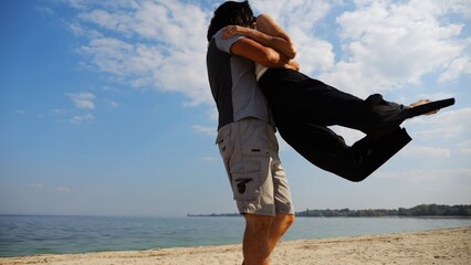 Happy couple having fun together on seashore at summer vacation. Man taking woman in air and turning around at coastline. Carefree lovers spending time at sandy beach. Concept of love. Slow motion
