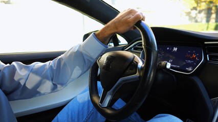 Male hand of businessman holds steering wheel while driving electrical vehicle at city. Businessperson operating electric car on urban road. Young man commuting to work at sunny day. Close up
