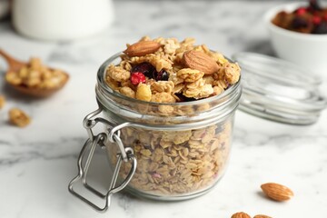 Tasty granola with dried fruits in glass jar on white marble table, closeup