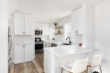 A white kitchen with hardwood flooring, decorations on floating shelves, and chairs sitting at the marble countertop.