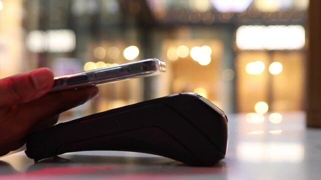 Close up of a customer making a mobile payment with a smartphone by tapping it on a pos terminal in a store, with a blurred background of a shopping mall showing bokeh lights