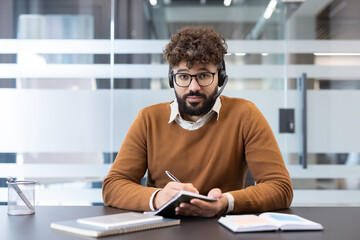 Young man wearing headset and glasses, working as a customer support consultant or attending a webinar, looking at camera and writing notes in an office setting
