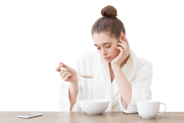 Portrait of young bored woman eating cereals with bad taste in her white loft kitchen, sitting at wooden table