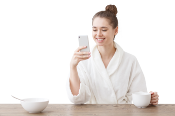 Horizontal picture of young European Caucasian lady wearing white bathrobe in morning eating breakfast and drinking tea or coffee at same time checking mail or chatting on cellphone, smiling
