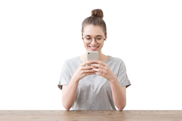 Happy young woman with her hair in a bun and glasses smiles while looking at her phone, sitting at wooden table