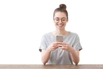 Happy girl with her hair in a bun and glasses smiles while looking at her phone, sitting at wooden table