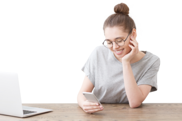 Smiling woman in glasses sitting at a wooden table looking at her phone with a laptop nearby