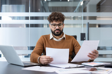 Young man analyzing important business documents and financial reports while concentrating at his desk in a modern corporate office, symbolizing hard work and analysis