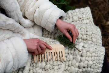 Woman in white coat holding pine twig and wooden comb on knit blanket