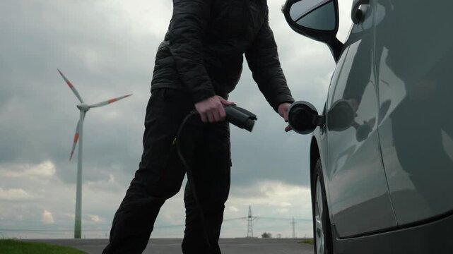 Unrecognizable person disconnects power connector from EV car, wind generator charging station. Man unplugs charging cable from electric vehicle near sustainable electricity, windmill, wind turbine