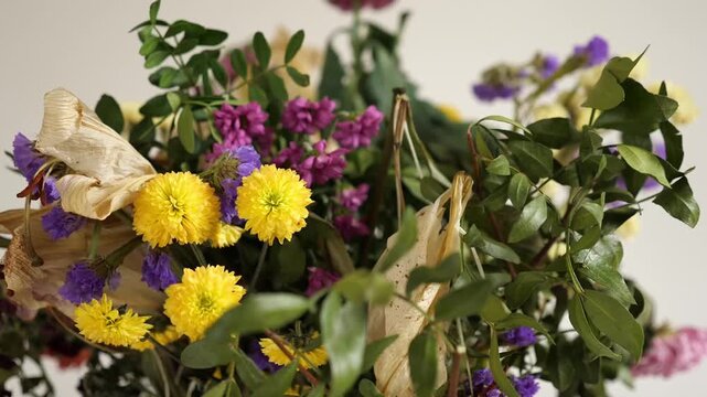A close-up horizontal indoor shot of a dried flower bouquet with purple Statice
(sea lavender), yellow globe amaranth, partially desiccated white lily, and green
foliage against a plain, light backgro