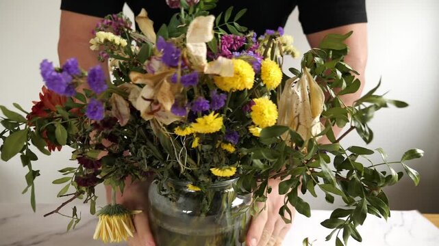 A close-up horizontal indoor shot of a dried flower bouquet with purple Statice
(sea lavender), yellow globe amaranth, partially desiccated white lily, and green
foliage against a plain, light backgro