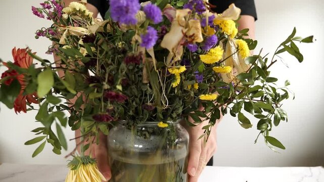 A close-up horizontal indoor shot of a dried flower bouquet with purple Statice
(sea lavender), yellow globe amaranth, partially desiccated white lily, and green
foliage against a plain, light backgro