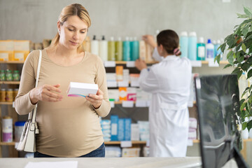 Fototapeta premium Pregnant woman studies composition of vitamins tablets in detail and buys useful supplements. Health care, healthy lifestyle, prevention. Girl pharmacist arrange showcase in background.
