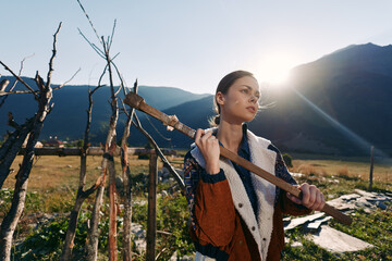 Naklejka premium woman countryside mountains farming field outdoors portrait sun backlit young farmer carrying wooden stick in rustic landscape, thoughtful profile wearing warm jacket and sweater at sunset.