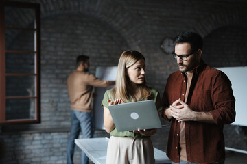 Two diverse colleagues discussing project while looking at laptop screen in modern startup office