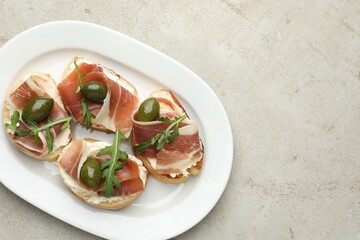 Delicious bruschettas with cream cheese, prosciutto, green olives and arugula on gray textured table, top view. Space for text