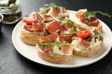 Delicious bruschettas with cream cheese, prosciutto, tomatoes and arugula on dark table, closeup