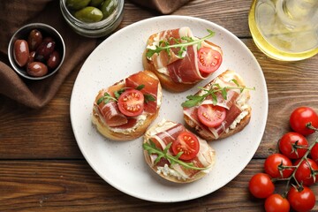 Delicious bruschettas with cream cheese, prosciutto, tomatoes, arugula and olives on wooden table, flat lay