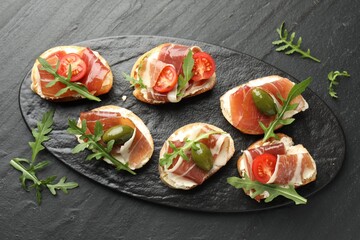 Delicious bruschettas with cream cheese, prosciutto, green olives, tomatoes and arugula on dark textured table, top view