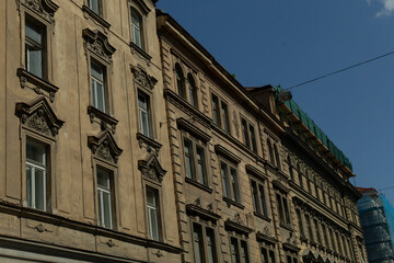 Obraz premium ornate historic facade under clear sky with sculpted pediments, tall sash windows, carved cornices and weathered stucco, warm stone patina, rooftop scaffolding and subtle urban skyline in background