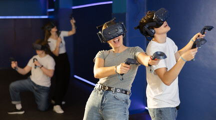Two teenagers in virtual reality glasses with joysticks stand shoulder to shoulder and play survival online game in stone jungle of future. concept of altered reality. Immersive technologies XR VR AR