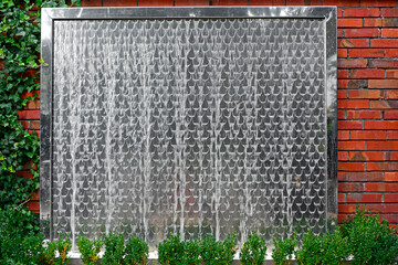 ściana wodna w ogrodzie. Metalowa ściana wodna na ceglanym murze. water wall in the garden, decorative water fountain cascading in garden, steel wall with a waterfall. Artificial decorative waterfall.