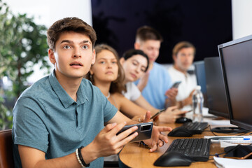 Excited young developer working at IT startup, watching project presentation with team gathered around table and using mobile phones for real-time data sharing