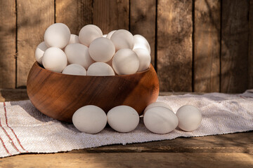 Chicken eggs in wooden bowl on a home-woven cloth, wall of chicken coop, barn or grunge fence in the background. Countryside outdoor setting in the backyard © TSViPhoto
