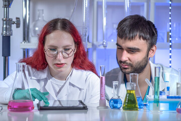 Young female lab technician working on a tablet while collaborating with male colleague in modern laboratory. Digital technology in chemistry research, teamwork, analysis and innovation concept.