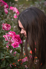 Naklejka premium Young woman smells pink roses in a garden, close-up portrait, soft daylight, rich green and pink tones, gentle and contemplative mood
