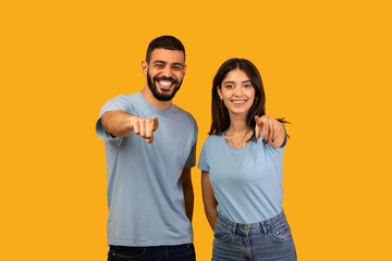 Obraz premium A young Middle Eastern couple stands against a bright yellow background. They are smiling and playfully pointing fingers at the camera, showing excitement and joy in their expression.