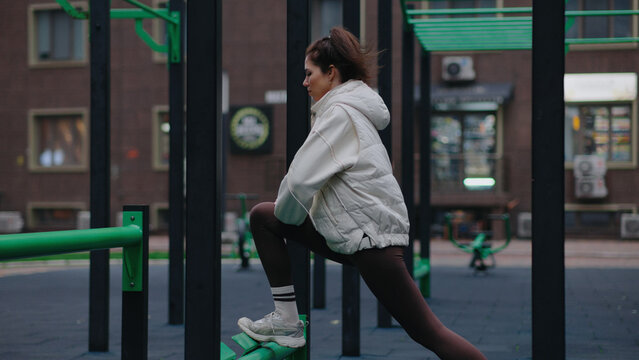 Young woman stretches at a fitness park in the afternoon sun while preparing for a workout session with calisthenics equipment nearby