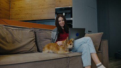 Woman sits on couch with laptop while petting small dog in a modern living room setting during daylight hours