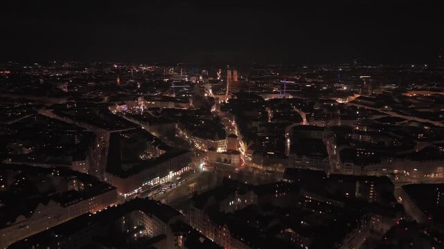 Drohnenperspektive bei Nacht ueber Muenchens Altstadt mit klar sichtbarer Frauenkirche, leuchtendem Neuen Rathaus, hell glitzerndem Weihnachtsbaum und dem Weihnachtsmarkt im Herzen der Stadt