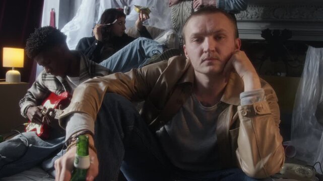 Front view shot of young man sitting on floor in abandoned house and looking at camera holding beer bottle with group of people in background, copy space