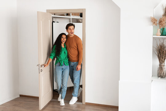 Happy young couple walks through the doorway of their new apartment, excitedly admiring the modern interior design. They share an emotional moment in their new home.