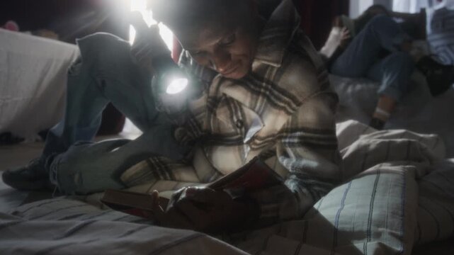 Handheld close up shot of African American man reading book with flashlight while lying on mattress in secret hideout of abandoned house, copy space