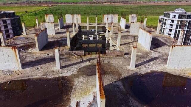 Concrete pillars rise from a vast, desolate landscape, remnants of a halted project contrasting with bright fields in the distance under a clear sky.